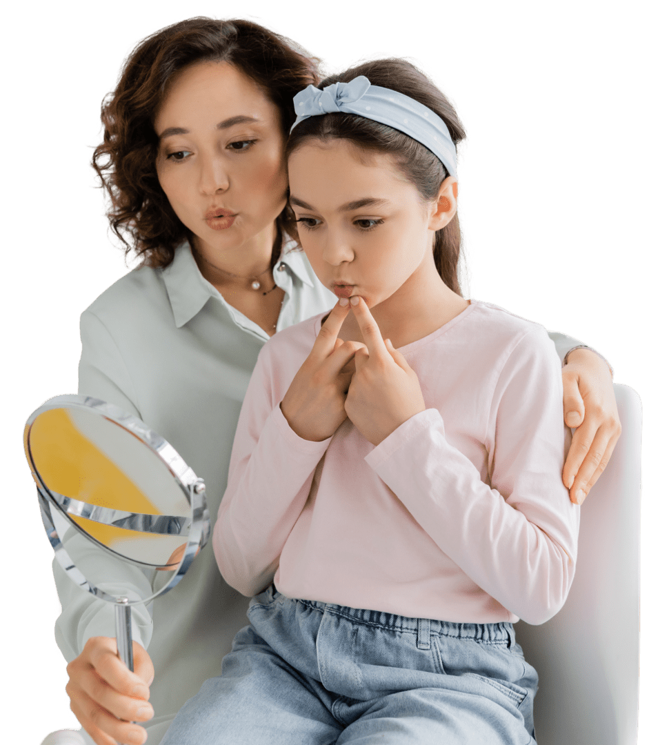 GettyImages-2182120916 1 A woman and a girl are sitting together, both exploring expressions in a hand mirror as part of speech therapy for children. The girl, wearing a headband and light pink shirt, joyfully mimics each expression.