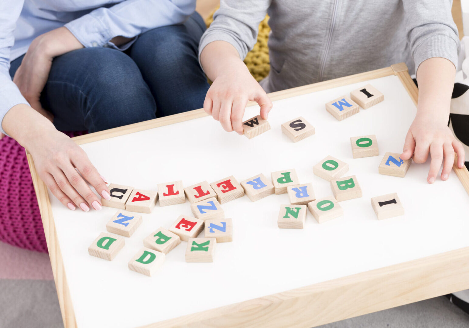 Two people sitting at a white table arranging alphabet blocks.